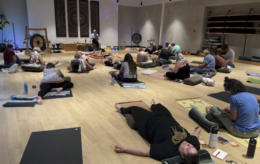 A group resting in restorative postures on mats and bolsters during a sound bath at Flow Yoga North Loop, with gongs at the front of the studio.