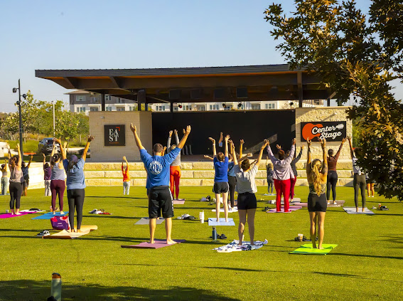 A group of people flowing with arms raised on the lawn in front of the Center Stage pavilion at EastVillage in Austin on a sunny morning.